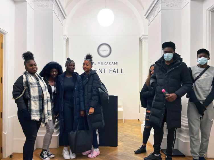 Our 15+ group standing inside of the entrance of the Superblue Exhibition. Background is white walls, with a round glowing white light in the top centre behind them. 